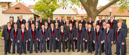 Ein Chor älterer Männer in formeller Kleidung, die vor einem Baum stehen, mit Gebäuden und Grün im Hintergrund.