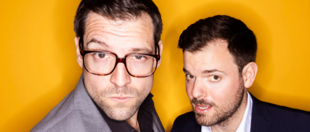 Zwei Männer posieren für ein Foto vor gelbem Hintergrund. Einer trägt eine Brille und einen grauen Anzug, der andere trägt einen schwarzen Anzug und ein weißes Hemd.