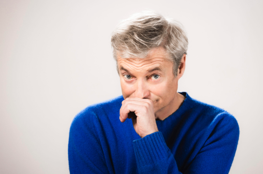 Ein Mann mit grauem Haar trägt einen blauen Pullover und hat seine rechte Hand am Mund. Er schaut nach rechts und scheint nachzudenken.