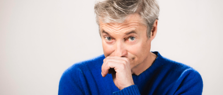 Ein Mann mit grauem Haar trägt einen blauen Pullover und hat seine rechte Hand am Mund. Er schaut nach rechts und scheint nachzudenken.