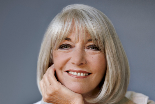 Eine ältere Frau mit kurzen blonden Haaren, lächelt mit der Hand auf dem Kinn.