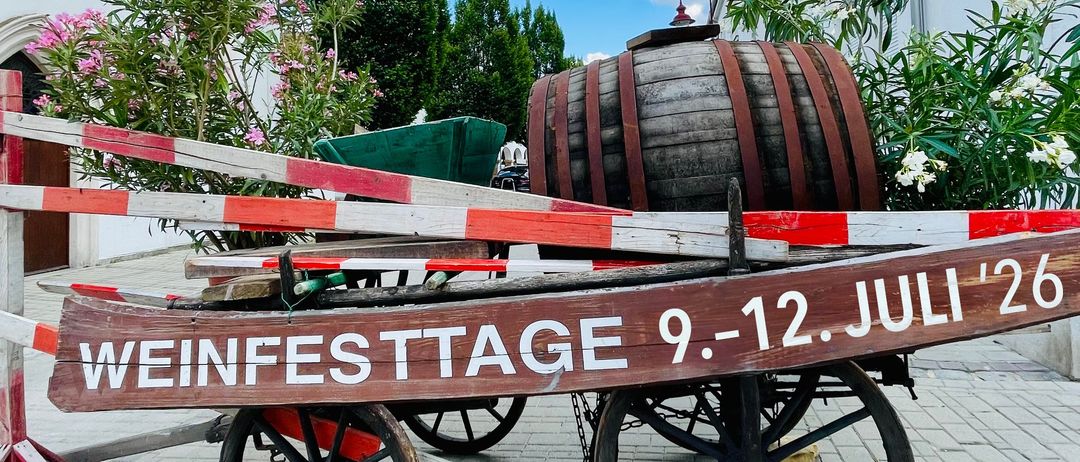 Ein Holzwagen mit einem Fass und einem Schild mit der Aufschrift Weinfesttage 9-12 Juli '26, vor einem Gebäude mit blauem Himmel im Hintergrund geparkt.