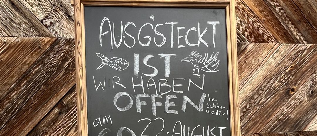 Ein Kreidetafel mit hölzerne Rahmen zeigt eine Nachricht auf Deutsch. Es steht 'AUSGSTECKT WIR HABEN OFFEN' mit Daten '22. August' und '23. August'. Darunter steht '5. SEPTEMBER' und 'RESERVIERUNG'. Eine E-Mail-Adresse 'fischundvogel@tav.at' ist ebenfalls geschrieben.