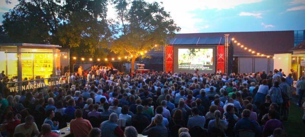 Eine große Menschenmenge im Freien sieht einen Film auf einer Leinwand. Viele Menschen sitzen an Tischen unter Lichterketten. Die Umgebung ist von Bäumen und Gebäuden umgeben.