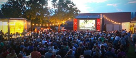 Eine große Menschenmenge sieht einen Film auf einer Leinwand im Freiluftkino. Tische und Stühle sind aufgestellt, und auf den Tischen stehen Flaschen und Getränke.