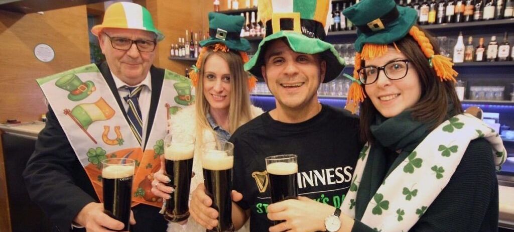 Vier Personen mit grünen Mützen mit orangefarbenen Bändern und Kleeblatt-Schals lächeln und halten Gläser mit Guinness-Bier in die Höhe. Sie stehen in einer Bar.