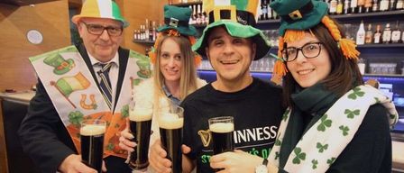 Vier Personen mit grünen Mützen mit orangefarbenen Bändern und Kleeblatt-Schals lächeln und halten Gläser mit Guinness-Bier in die Höhe. Sie stehen in einer Bar.