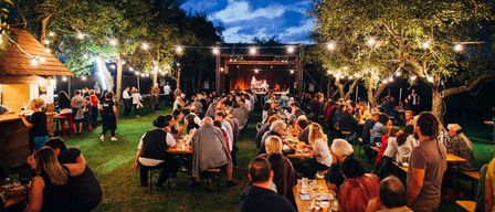 Ein nächtliches Outdoor-Essevent mit vielen Gästen, die an Tischen unter Lichterketten sitzen. In der Ferne steht eine Bühne mit Künstlern.