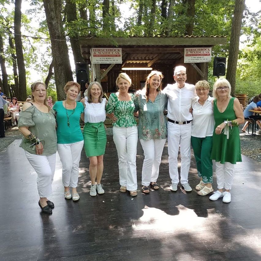 Eine Gruppe von Erwachsenen, gekleidet in Weiß und Grün, posiert für ein Foto vor einer Holzstruktur mit Schildern, die Hirschrock und Waldela lesen.