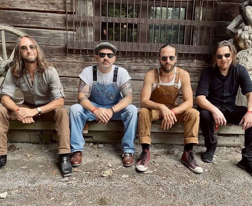 Fünf Männer in Sonnenbrillen und Latzhosen sitzen auf einer Holzbank vor einem Holzgebäude.