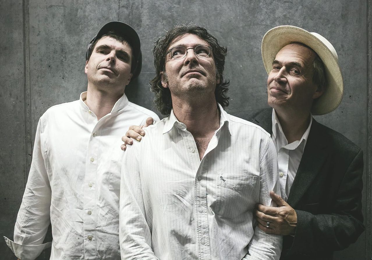 Three men in white shirts stand together, one wearing a hat, another with glasses, and the third smiling. They appear to be posing for a photograph.
