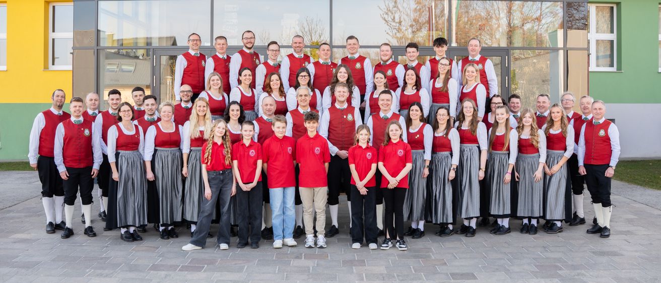 Ein Chor, einige in Rot und andere in Weiß und Grau gekleidet, posiert vor einem Gebäude mit Glaswänden.