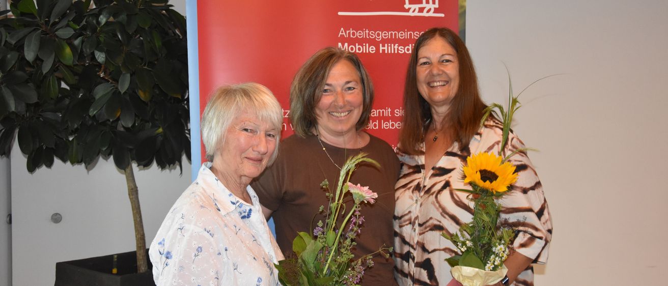 Drei Frauen stehen nebeneinander und lächeln. Sie halten Blumensträuße und stehen vor einem roten Banner.
