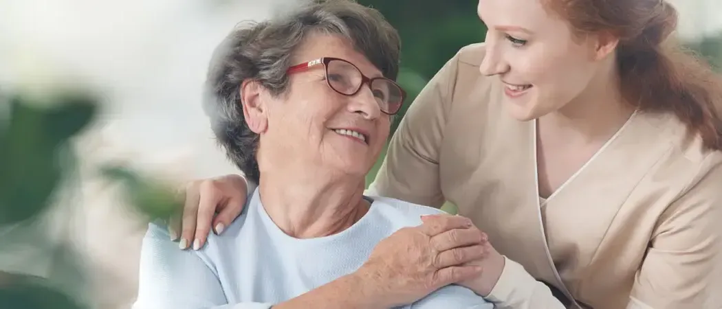 An elderly woman with glasses smiles while a young woman hugs her shoulder outside.
