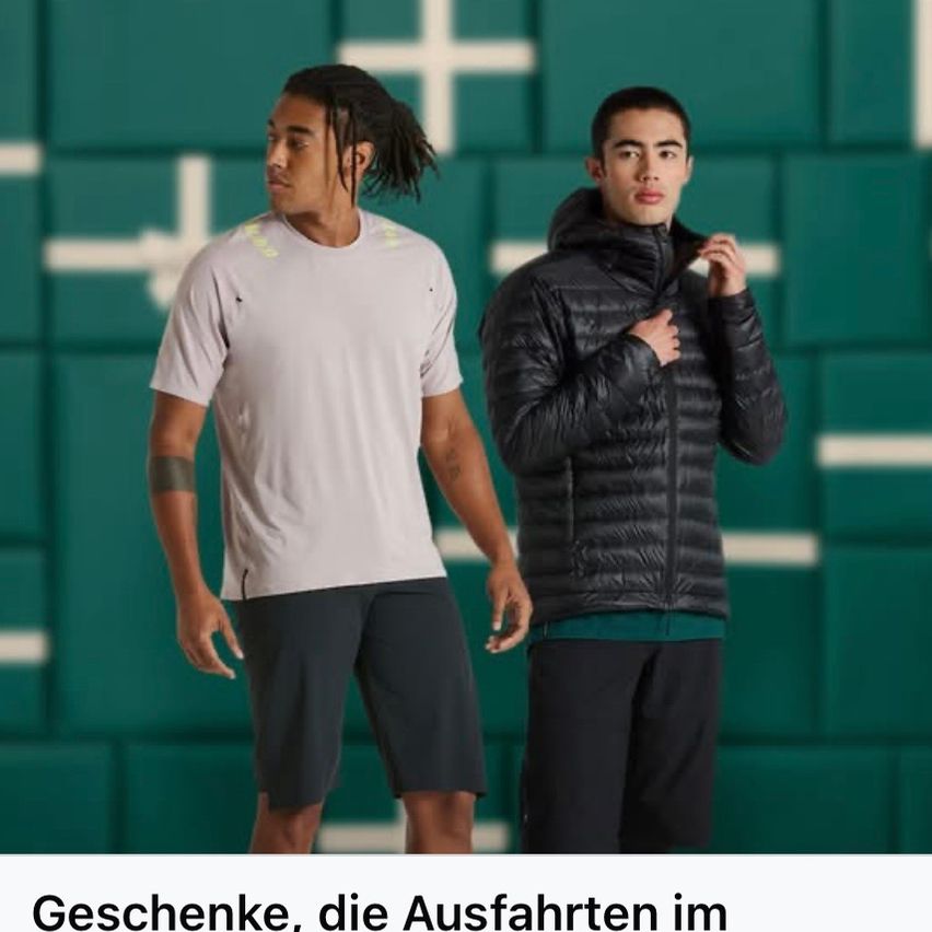 Zwei Männer stehen nebeneinander vor einer grünen Wand mit weißen Kreuzen. Ein Mann hat einen Zopf und trägt ein weißes T-Shirt und schwarze Shorts. Der andere Mann hat eine schwarze Daunenjacke und schwarze Hosen.