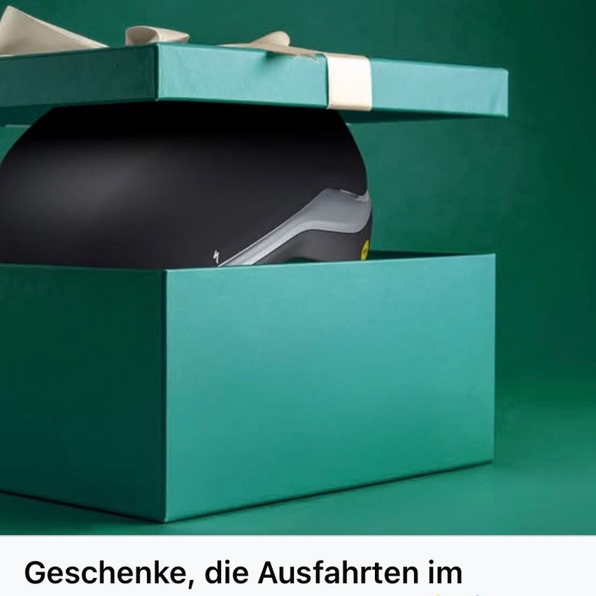 Eine grüne Box mit einem weißen Band und einer Schleife enthält einen schwarzen Helm. Die Box hat einen Deckel und der Helm ist darin platziert.