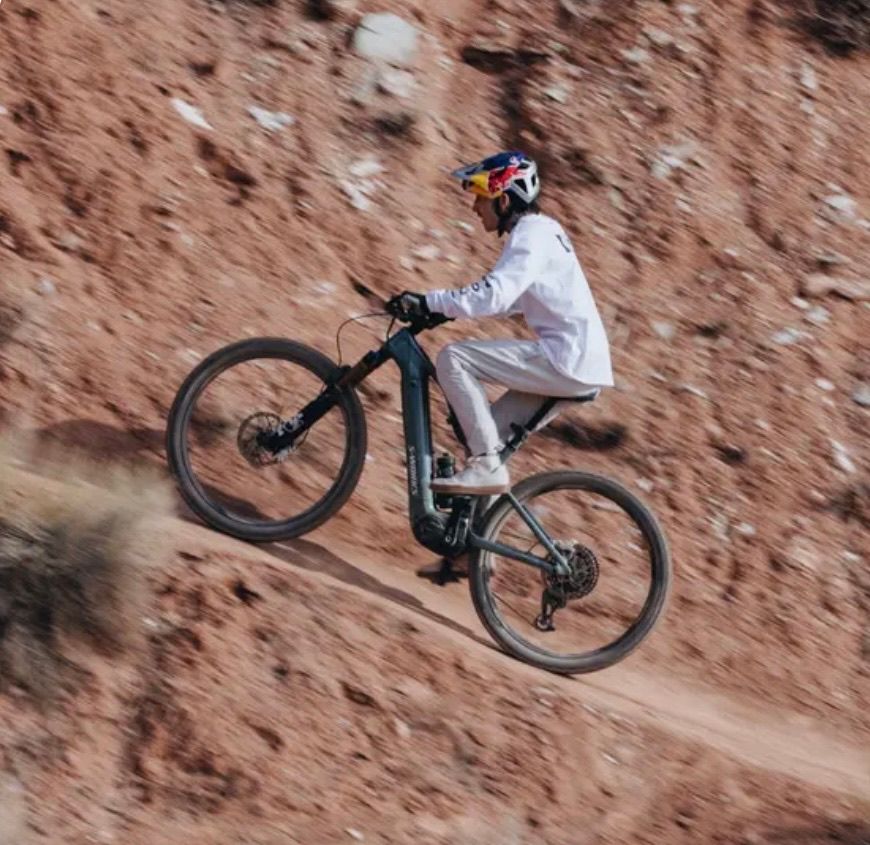 A person rides a black electric mountain bike on a steep dirt trail. The rider wears a helmet, gloves, and sneakers. The scene is set in a rugged, arid landscape.