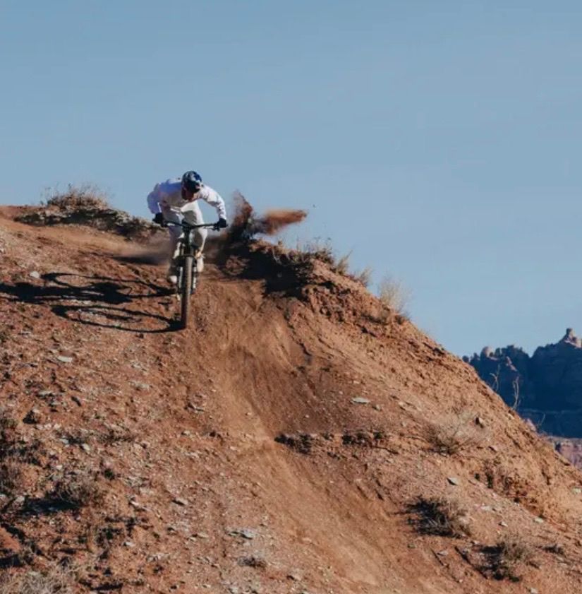 A person rides a mountain bike down a steep, dirt trail with scattered rocks under a clear blue sky.