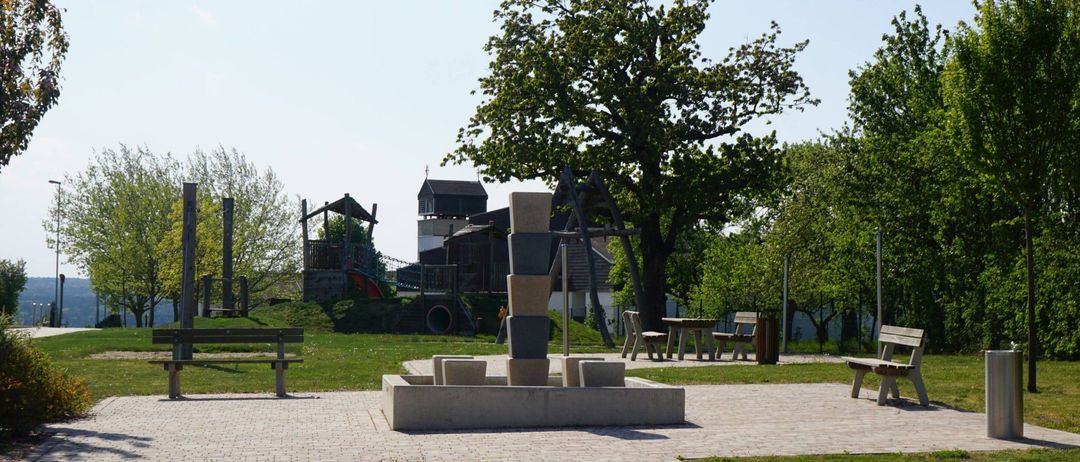 Ein Park mit einem Ziegelweg, Bänken und einem kleinen Brunnen. Im Hintergrund befinden sich Bäume und ein Spielplatz. Der Himmel ist klar.
