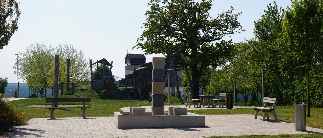 Ein Park mit einem Ziegelweg, Bänken und einem kleinen Brunnen. Im Hintergrund befinden sich Bäume und ein Spielplatz. Der Himmel ist klar.