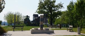 Ein Park mit einem Ziegelweg, Bänken und einem kleinen Brunnen. Im Hintergrund befinden sich Bäume und ein Spielplatz. Der Himmel ist klar.
