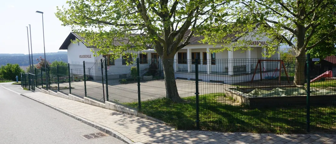Ein weisses Gebäude mit braunem Dach, beschriftet mit Volksschule, ist von einem grünen Zaun umgeben. Davor steht ein Baum und ein Spielplatz mit Schaukel.