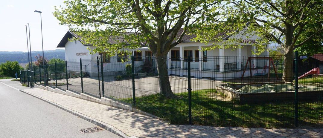 Ein weisses Gebäude mit braunem Dach, beschriftet mit Volksschule, ist von einem grünen Zaun umgeben. Davor steht ein Baum und ein Spielplatz mit Schaukel.