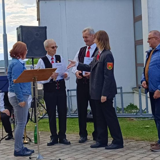 Eine Gruppe von Menschen steht auf einem Ziegelboden. Ein Mann in einem Anzug überreicht einer Frau in einer Blazerjacke ein Papier. Ein Mann in einer Weste hält ein Papier. Eine Frau in einer Jeansjacke schaut sie an. Ein Lautsprecher steht hinter ihnen. Ein Metallzaun und ein Gebäude sind hinter ihnen.