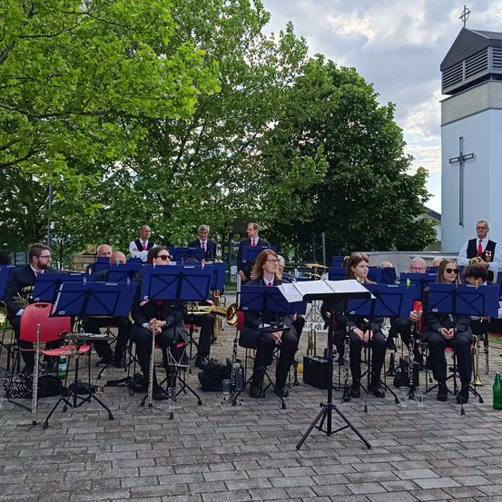 Eine Band in formeller Kleidung spielt Instrumente auf einem gepflasterten Weg, mit Bäumen und einer Kirche im Hintergrund.