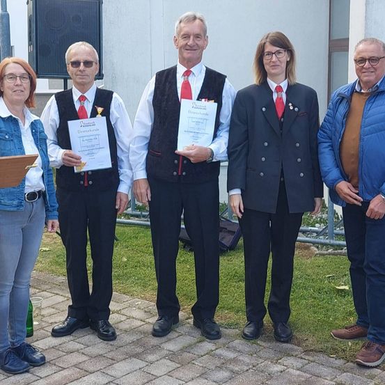 Fünf Personen stehen auf einem gepflasterten Weg, wahrscheinlich bei einer Veranstaltung. Sie lächeln alle und scheinen für ein Foto zu posieren. Jedes hält eine Urkunde in der Hand, außer der Frau auf der linken Seite, die eine Mappe hält. Der Mann auf der rechten Seite trägt eine blaue Jacke, während die anderen formelle Kleidung tragen.