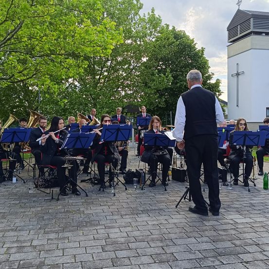 Eine Band spielt auf einer gepflasterten Fläche mit einem kreuzbedeckten Gebäude im Hintergrund. Einige Mitglieder sitzen, andere stehen, und sie spielen verschiedene Instrumente.