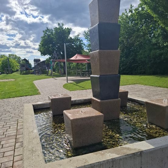 Ein Betonbrunnen mit einem Stapel von Blöcken steht in einem Park mit grünem Rasen und Bäumen. In der Ferne befindet sich ein Spielplatz mit einem roten Baldachin.