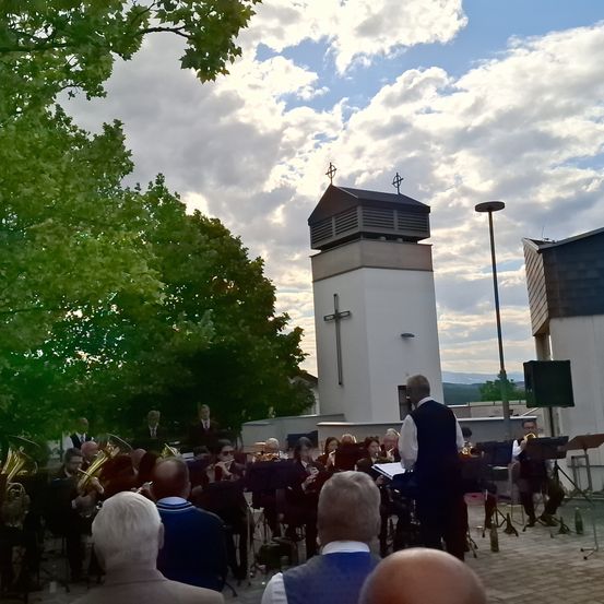 Ein Orchester spielt auf einer Bühne im Freien mit Bäumen und einer Kirche im Hintergrund. Das Publikum sitzt vor ihnen.