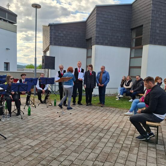 Eine Band spielt vor einem modernen Gebäude. Musiker spielen Instrumente, und Zuschauer sitzen auf Bänken. Einige Leute stehen davor, darunter eine Frau mit einem Papier. Der Himmel ist bewölkt.