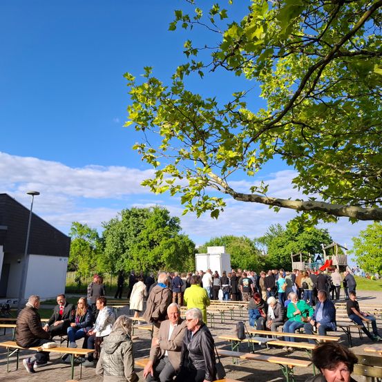 Eine Gruppe von Menschen sitzt auf einer großen Freiluft-Picknickfläche mit Holzbänken. Bäume und ein Gebäude sind im Hintergrund zu sehen, und der Himmel ist blau mit einigen Wolken.