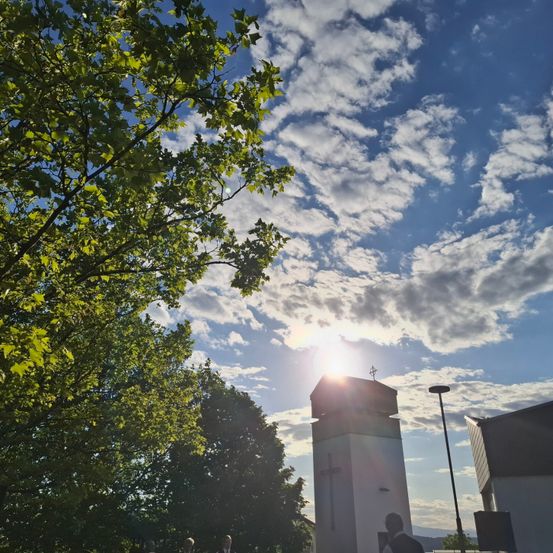 Ein sonniger Tag mit einem klaren blauen Himmel, der von Wolken durchsetzt ist, einem hohen weißen Turm mit einem Kreuz oben und mehreren Menschen, die darunter gehen.