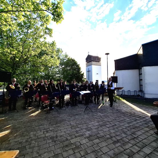 Eine Musikergruppe spielt im Freien, umgeben von Bäumen und einem Gebäude. Sie spielen verschiedene Instrumente auf einem gepflasterten Boden.