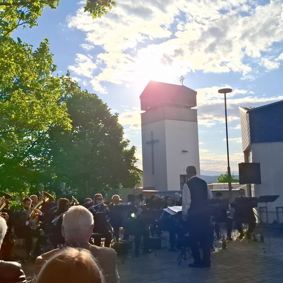 Eine Brassband spielt im Freien vor einer Kirche. Ein Dirigent steht vor den Musikern, die sitzen. Bäume und ein Turm sind im Hintergrund.