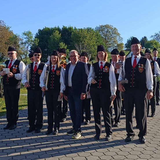 Eine Gruppe von Musikern in traditioneller Kleidung, die in einer Reihe stehen, Instrumente halten und für die Kamera lächeln. Bäume und ein Haus sind im Hintergrund.