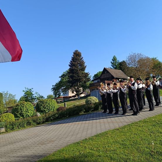 Ein Blasorchester in traditioneller Kleidung steht auf einem gepflasterten Weg und spielt vor einem Haus und Bäumen an einem sonnigen Tag.