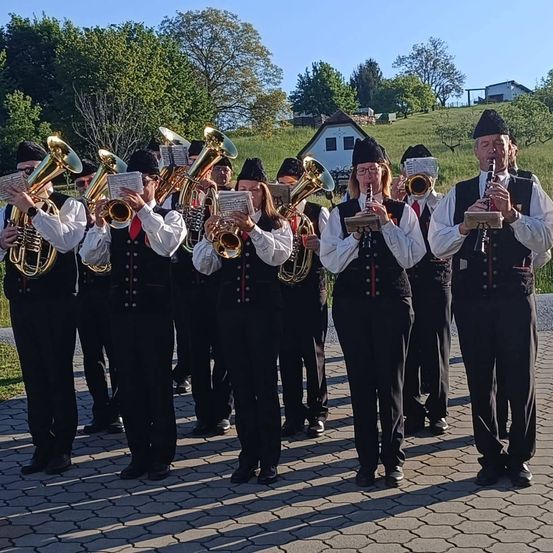 Eine Blaskapelle steht auf einem gepflasterten Gehweg, hält Instrumente in den Händen und trägt traditionelle Uniformen. Sie sind von Grünflächen und einem Haus im Hintergrund umgeben.