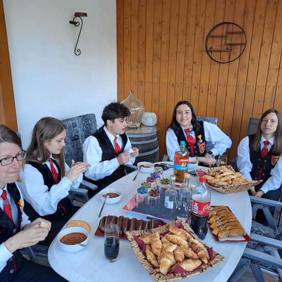 Fünf Personen, gekleidet in identischen Uniformen, sitzen um einen Tisch herum. Der Tisch ist mit verschiedenen Speisen bedeckt, darunter Brötchen, Desserts und eine Flasche Coca-Cola. Eine Frau auf der linken Seite hält ein Stück Brot. Hinter ihnen ist eine Holzwand mit einem dekorativen runden Objekt zu sehen.