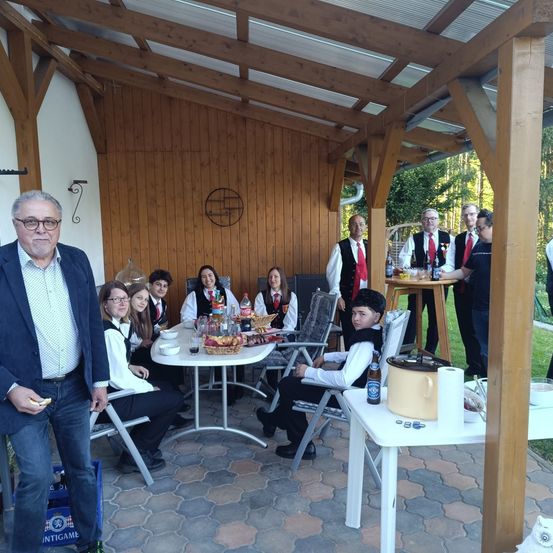 Eine Gruppe von Menschen in traditioneller Kleidung, die um einen Tisch mit Essen, Getränken und einer Flasche sitzen, mit einem hölzernen Pavillon im Hintergrund.