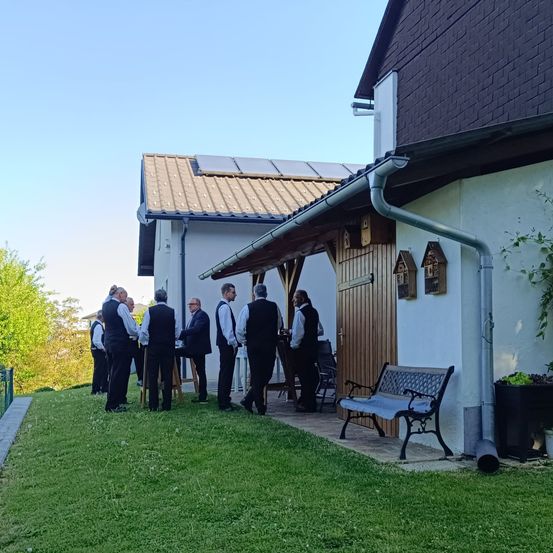 Mehrere Männer in formeller Kleidung stehen auf der Veranda eines Hauses mit Solarmodulen. Sie scheinen sich zu unterhalten. Eine Bank und ein Topfpflanzen sind in der Nähe.
