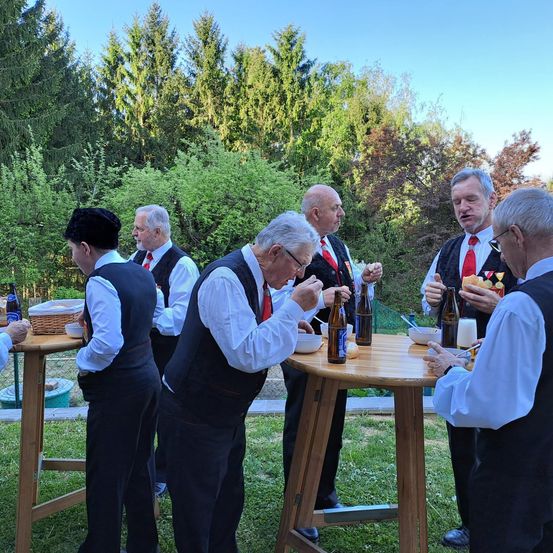 Eine Gruppe älterer Männer steht um einen Tisch herum, isst und trinkt. Sie sind in formeller Kleidung gekleidet. Der Tisch ist mit Flaschen, Schüsseln und Brot beladen. Die Szene spielt sich im Freien ab, mit Bäumen im Hintergrund.