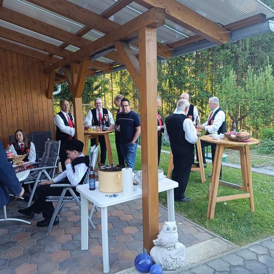 Mehrere Menschen versammeln sich in einem überdachten Außenbereich mit Holzstrukturen. Sie sind in verschiedenen Kleidungsstücken gekleidet, und einige sitzen auf Stühlen. Es gibt Tische mit Gegenständen darauf, und eine Person steht vor einem Tisch mit einer Schüssel.
