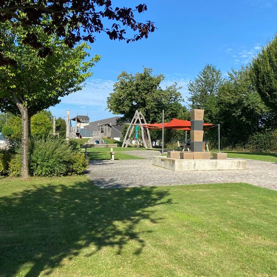 Ein Außenpark mit einem Betonbauwerk, einem roten Baldachin, einer dreieckigen Holzstruktur und Bäumen an einem sonnigen Tag.