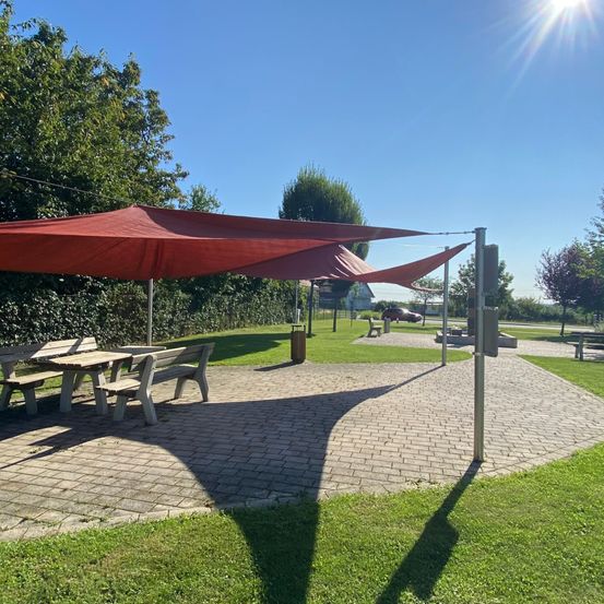 Ein Parkbereich mit einem großen roten Sonnensegel über einem Picknicktisch. Es gibt Bänke, einen Mülleimer und ein rotes Auto, das in der Ferne geparkt ist.