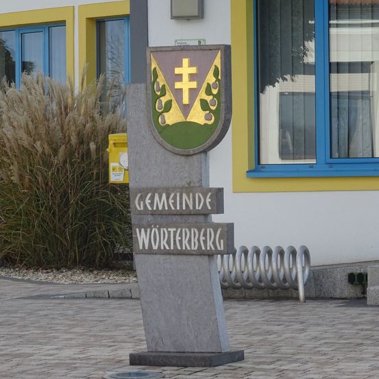 Ein Betonschild mit einem Wappen und dem Namen Gemeinde Worterberg steht vor einem Gebäude mit gelber und blauer Einfassung.