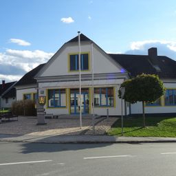 Ein weißes Gebäude mit gelber Umrandung hat mehrere Fenster und einen Fahnenmast vorn. Der Eingang befindet sich unter einem Vordach. Davor stehen Bänke und ein Baum.
